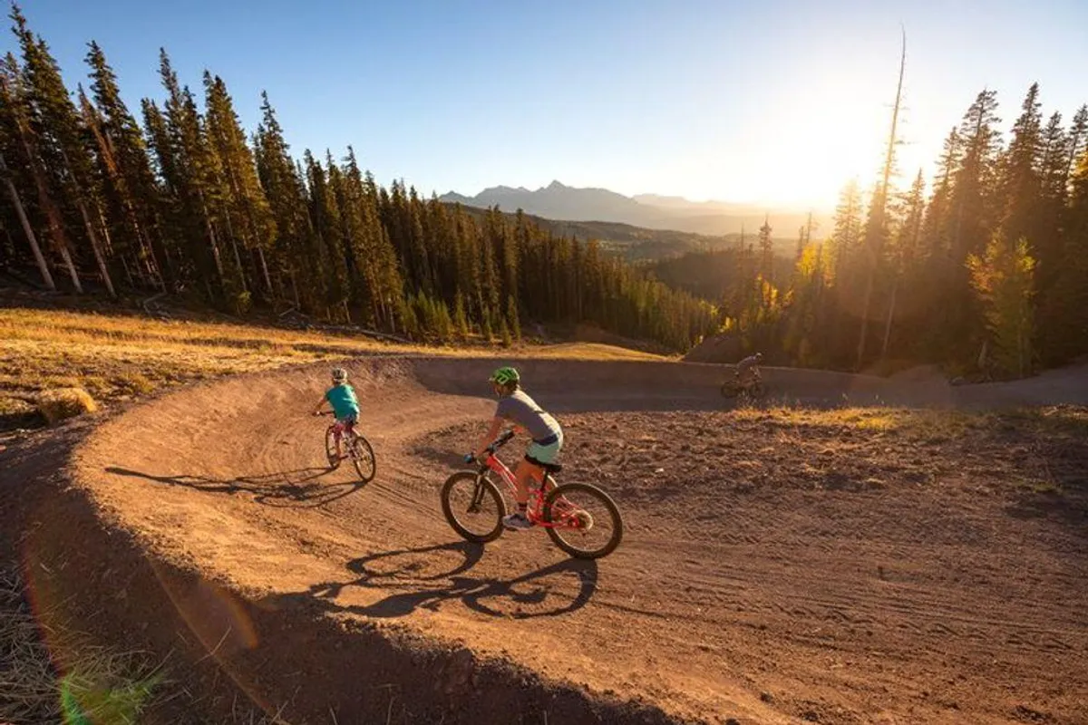 This exterior image captures a group of mountain bikers riding along a winding trail through a dense forest, illuminated by the golden glow of sunset. The scene is dynamic and vibrant, evoking the thrill and natural beauty of Telluride's mountain biking adventures.