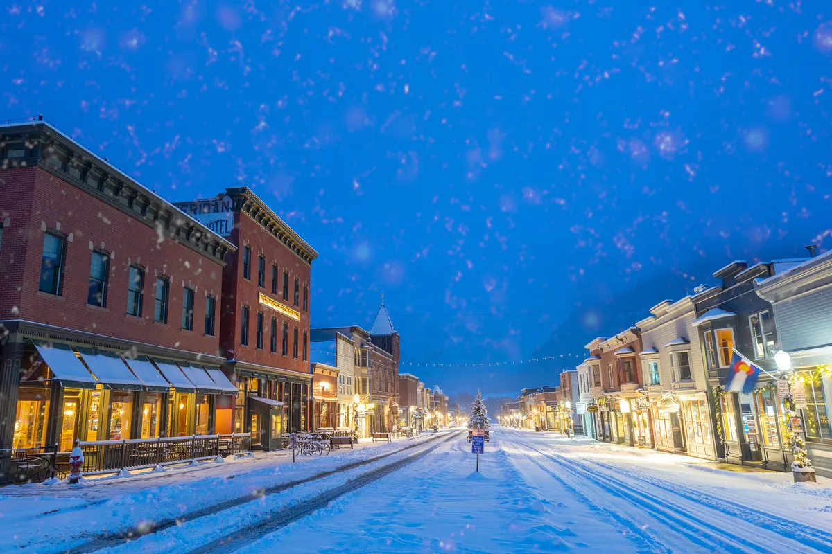 A snow-covered street lined with festively lit buildings, with a Christmas tree glowing in the distance. People stroll along the sidewalks, some carrying shopping bags, others sipping hot beverages at outdoor cafes. The overall ambiance is warm and inviting, despite the winter chill.