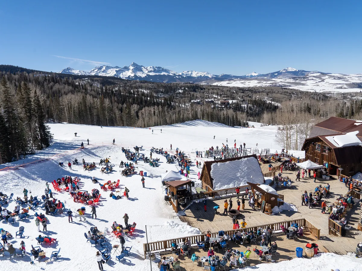 A dynamic ski area scene with people enjoying winter activities against a backdrop of snowy mountains. There are skiers and snowboarders, outdoor seating areas for après-ski relaxation, and wooden structures that add to the mountain resort vibe.
