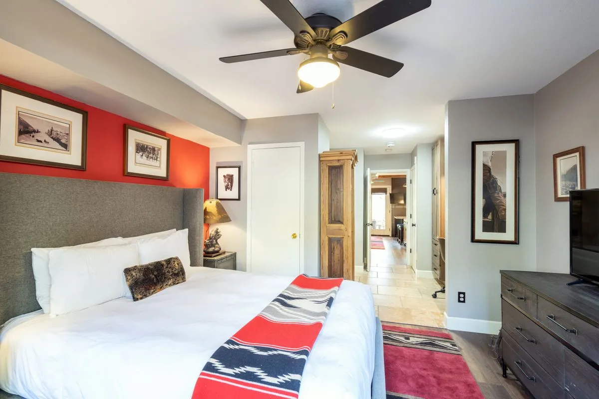 The image showcases a warm, inviting bedroom with a plush bed adorned with a decorative throw, tasteful wall art, a rustic wooden armoire, and a television—bathed in gentle natural light. The scene suggests comfort and relaxation, ideal for unwinding after a day of mountain adventures.