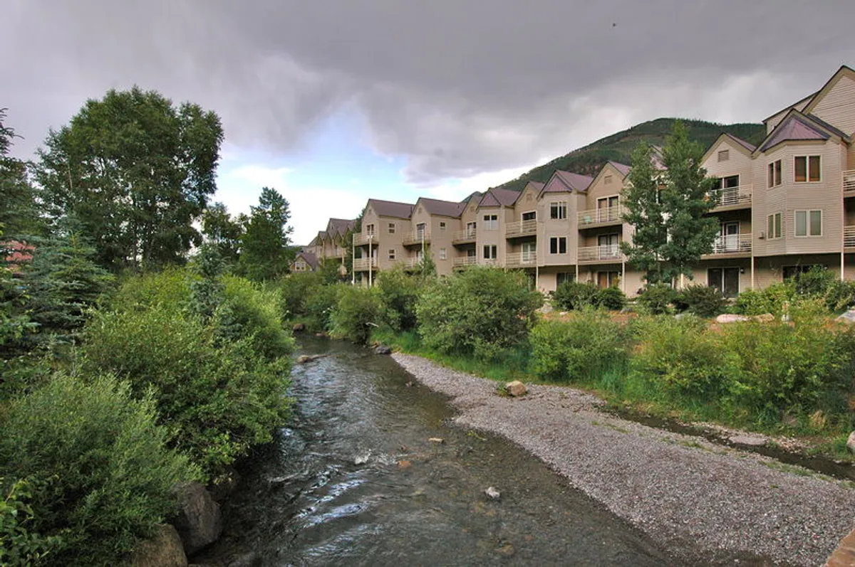 A serene riverside scene featuring the flowing stream, the buildings nestled beside the water, and mature trees providing shade. The view emphasizes the property’s direct riverfront location and visual connection to the River Trail.