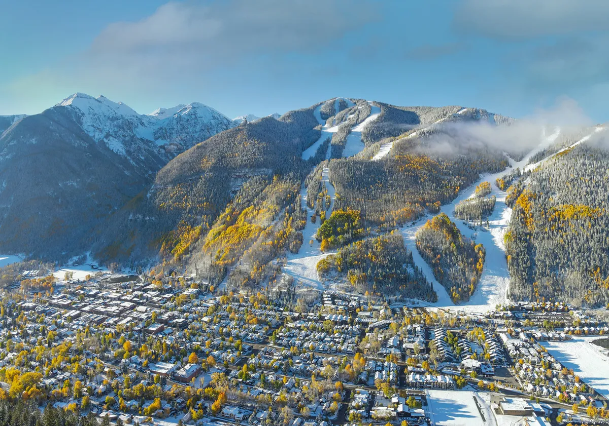 A panoramic view reveals snow-capped mountains, colorful autumn foliage, and the bustling town of Telluride nestled below. Ski slopes cut through the landscape, blending the seasons of fall and winter in one captivating scene.
