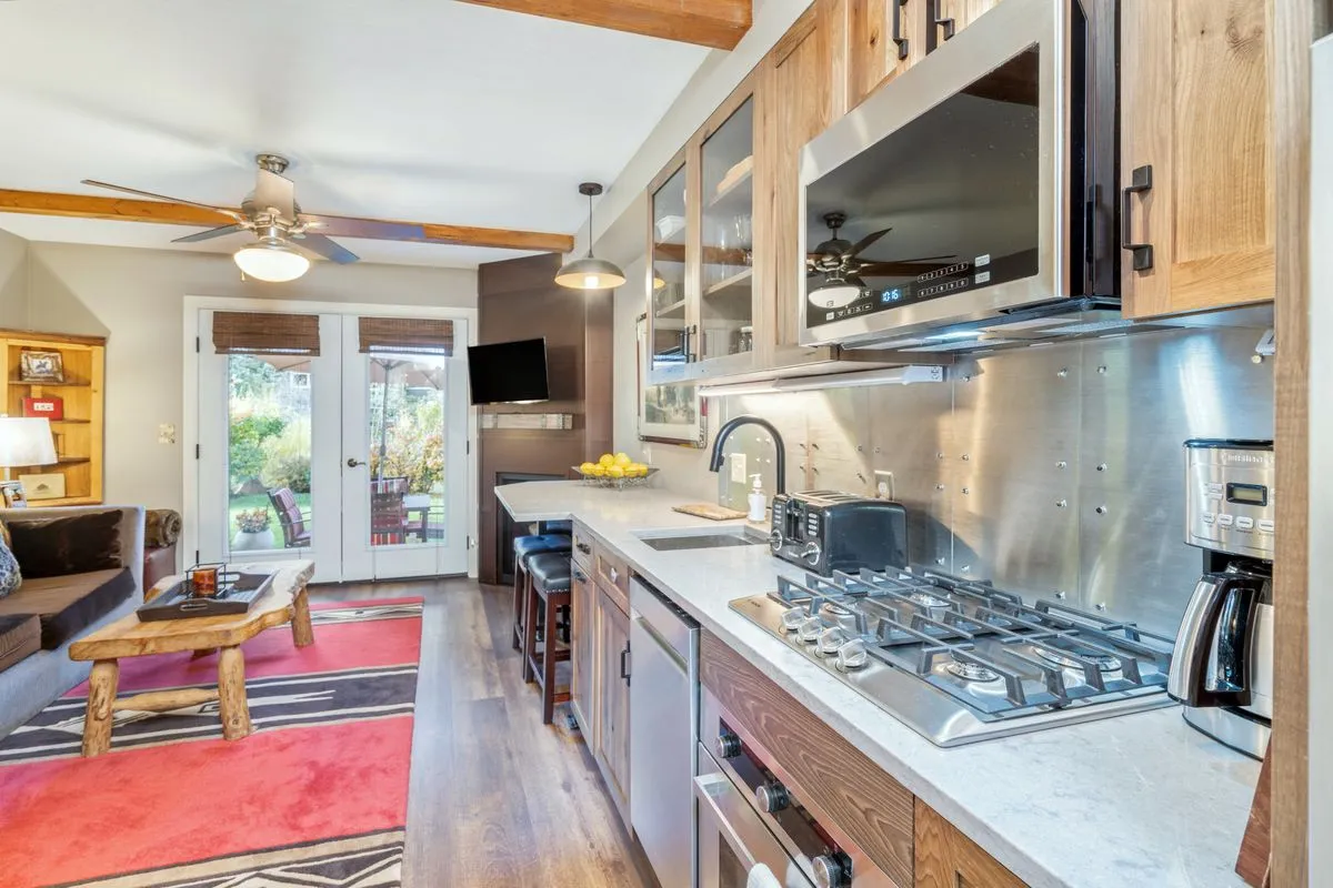 This image showcases a compact kitchenette with a central kitchen island, modern appliances (including a stove, oven, microwave, and refrigerator), and visible wooden ceiling beams. There's also a comfortable sofa with patterned throw pillows, reinforcing a homey, family-friendly atmosphere. The space is bright and functional, exemplifying the convenience highlighted in the section.
