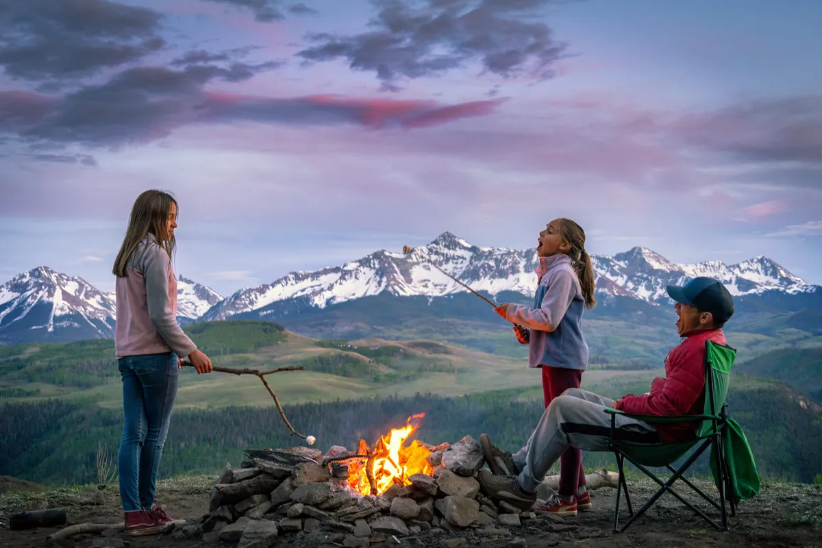 A warm, joyful outdoor evening around a campfire, with children roasting marshmallows and an adult relaxed in a chair. The background features a beautiful mountain landscape, evoking togetherness and outdoor enjoyment.