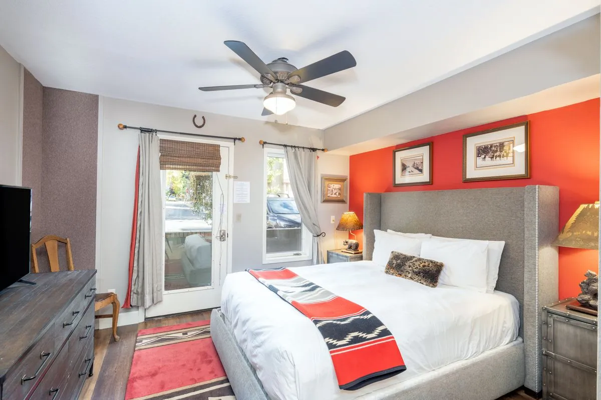 A bright bedroom area with a neatly made queen bed, a window letting in natural light, a dresser, lamp, and tasteful artwork on the wall. The room feels comfortable and welcoming, perfect for restful nights and lazy mornings.