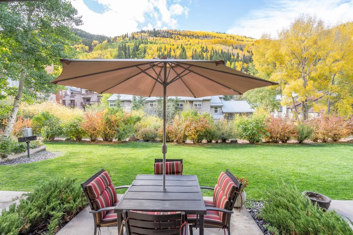 A private patio area with a table and umbrella surrounded by lush foliage, with expansive mountain views in the background. The patio setting is peaceful, inviting, and directly connected to natural surroundings, suggesting direct access to outdoor trails or the river nearby.