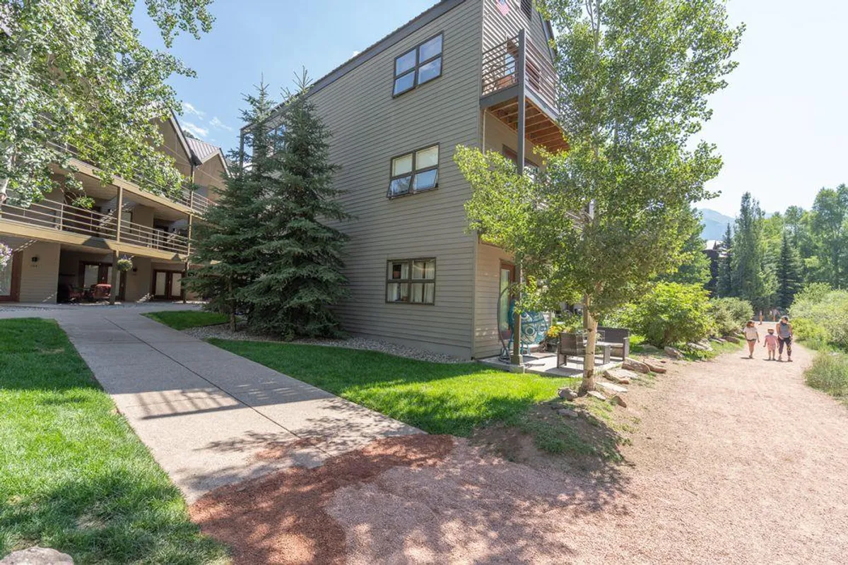 This image showcases the building's facade set against lush greenery and a walking path that leads directly into nature. People can be seen walking and relaxing outdoors, with the landscaping and mountain surroundings emphasizing the close connection to the natural environment.