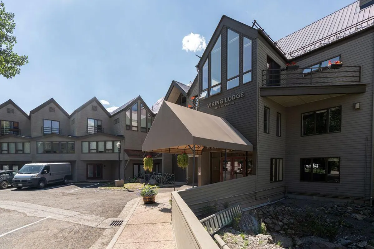 The building's welcoming entrance is visible, with large windows, bikes parked outside, and vibrant planters filled with flowers. The scene is lively and inviting, suggesting a space that encourages outdoor exploration and communal relaxation.