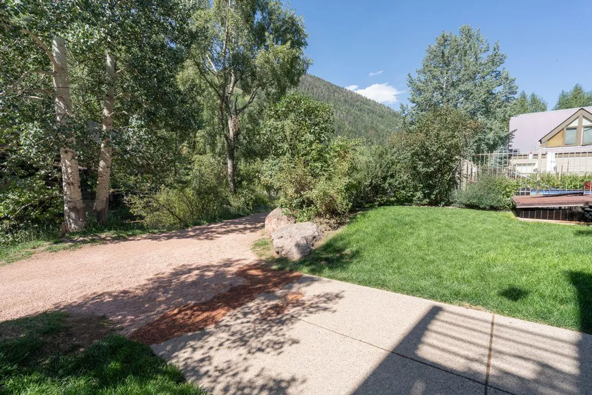 This image presents a tranquil pathway bordered by green grass and mature trees, with mountains in the background. It suggests peaceful walks, quiet moments, and the transition of seasons, making it ideal for morning coffees or nature observation.
