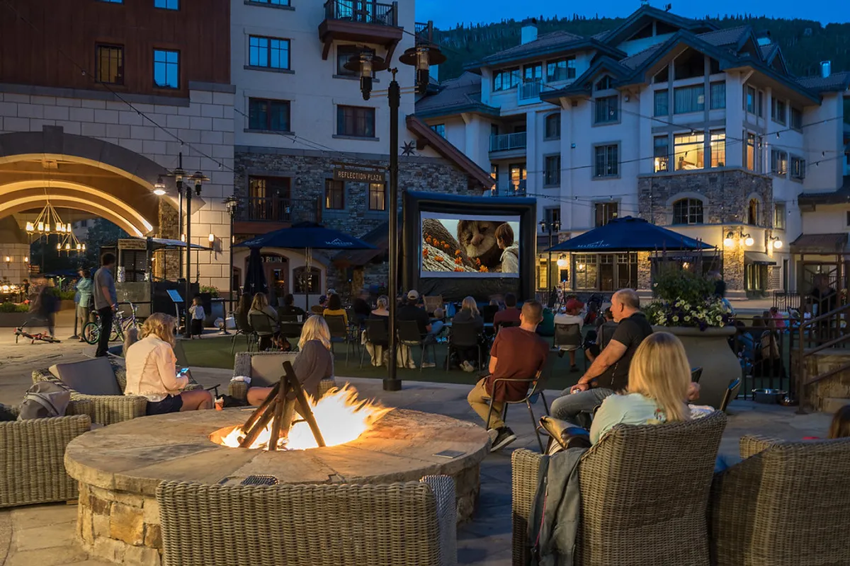 Guests gather around a glowing fire pit, watching a movie projected on an outdoor screen. The setting is cozy and communal, with people enjoying warm drinks and shared laughter under the night sky.