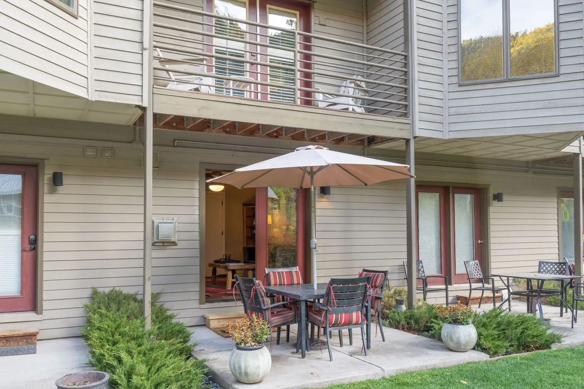 An outdoor dining area with a patio table, umbrella, decorative planters, and a balcony, set against a backdrop of greenery. The scene radiates warmth, inviting guests to relax outside, enjoy meals, or gather for conversations while soaking in the sunlight.