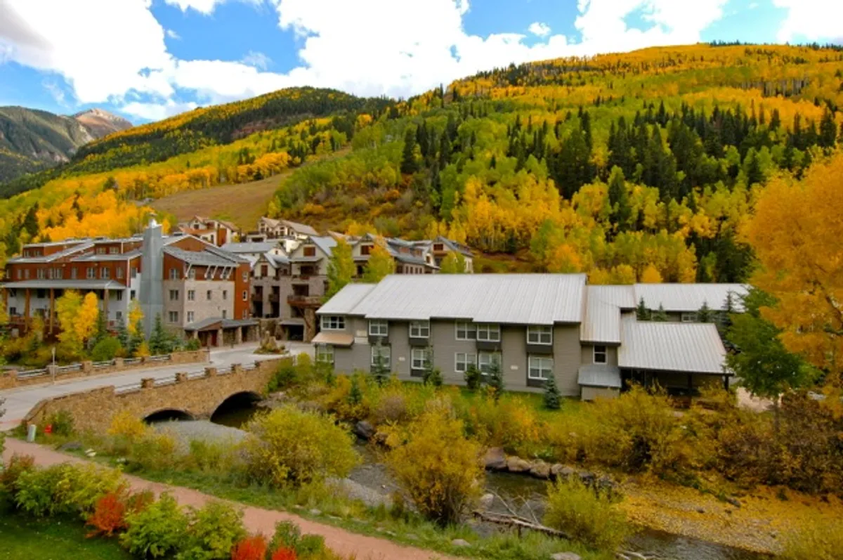A picturesque riverside scene featuring vibrant autumn foliage, a flowing stream, mountain backdrop, and quaint nearby buildings. The colors evoke the magic of fall, while the serene environment suggests peaceful strolls and moments of reflection.