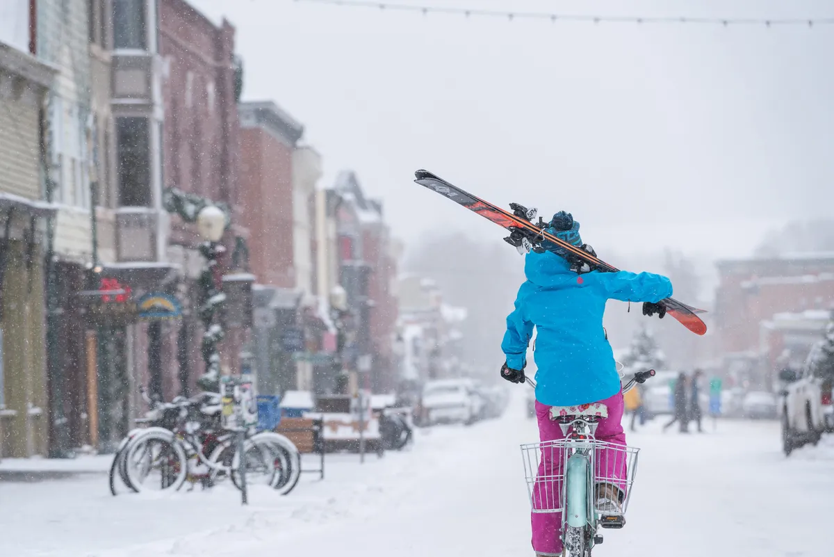 A lively winter street scene featuring a person holding skis high above their head, historic buildings dusted with snow, bicycles parked along the street, and people exploring shops. The mood is joyful and adventurous, capturing the dynamic energy of the town.
