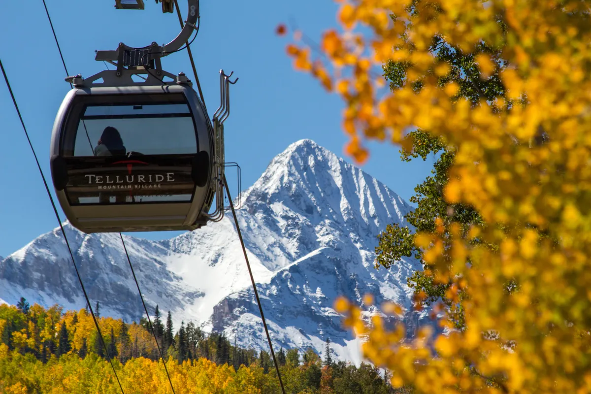 A gondola lift ascends towards a snow-capped mountain, surrounded by vibrant autumn foliage. The scene hints at year-round adventure, with opportunities for hiking, sightseeing, and exploring high mountain terrain.